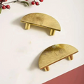 Two gold wall-mounted shelf hooks with a sunburst textured design and small wooden legs, displayed on a white wall with decorative red berries in the corner.