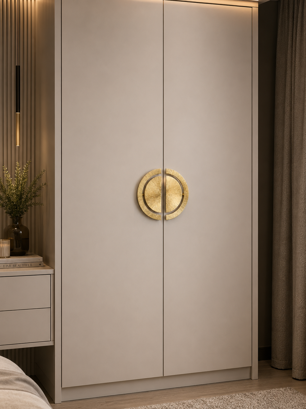 A modern two-door wardrobe or closet in a warm light beige finish, featuring a prominent round gold metal ring handle on the center of the doors, set in a styled bedroom next to a nightstand and curtains.